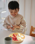 A young child in a striped shirt smiles while peeling an egg at a table set with mushie Round Dinnerware Plates, next to sliced apples and raspberries, as sunlight streams in through the window behind him.