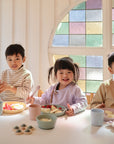 Three children sit at a table eating snacks, smiling in a bright room with a colorful stained glass window. mushie Round Dinnerware Plates, made of non-toxic plastic, are neatly arranged in front of them.