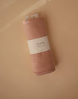 A rolled-up Mushie Ribbed Baby Blanket in peach, made from organic cotton, sits on a beige surface. The breathable blanket features a white Mushie label and two white tags at the top.