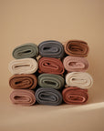 Twelve Mushie Ribbed Baby Blankets in beige, brown, pink, green, and gray are rolled and stacked in a 3x4 grid against a plain light tan background.