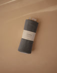 A rolled-up Ribbed Baby Blanket by Mushie in dark gray organic cotton rests on a light beige surface with soft shadows.
