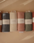 Six ribbed Mushie organic cotton baby blankets in earthy tones are lined up side by side. Each breathable blanket features a white Mushie label on a warm beige background.