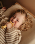 A laughing toddler with curly blonde hair lies in a crib, wearing a brown striped onesie and chewing on the mushie Flower Training Toothbrush.