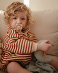 A young child sits on a beige couch in a striped outfit, holding a Mushie FRIGG Rope Natural Rubber Pacifier 2-Pack in their mouth and hugging a crescent moon pillow—true baby essentials.