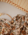 A mushie Organic Cotton Muslin Swaddle Blanket with a floral pattern and a beaded pacifier clip are placed on a white sheet inside a light wooden crib.