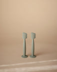 Two Mushie Diaper Cream Applicators in light green silicone stand upright on a beige background, highlighting their minimalistic design and practical use for applying diaper cream.