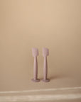 Two pastel purple Mushie Diaper Cream Applicators stand upright on a beige surface with a matching background. The minimalistic scene is softly lit, highlighting the silicone applicators' gentle design.