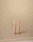 Two pale pink Mushie Diaper Cream Applicators, made of silicone and designed for applying diaper cream, stand upright side by side on a neutral beige surface with a plain beige background.