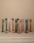 Thirteen minimalistic, abstract figures in muted pastels stand together on a beige background, their smooth forms inspired by the Mushie Diaper Cream Applicator.