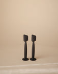 Two black Mushie Diaper Cream Applicators with flat, rectangular heads stand upright on a beige surface and background, resembling modern silicone applicators designed for easy, mess-free diaper cream application.