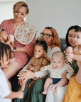 Four women sit together on a couch, each holding a baby using mushie Organic Cotton Muslin Burp Cloths, while a toddler stands nearby. The group is happy and engaged, filling the room with smiles and warmth.