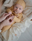 A baby in a mustard yellow outfit and hat rests in a cushioned bassinet with a floral blanket. Nearby, a mushie Organic Cotton Muslin Cloth from the 3-Pack lies close as an adult gently holds the baby's hands.