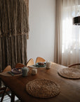 A cozy dining room showcases a wooden table set for breakfast with Mushie's Classic Silicone Suction Plate, woven placemats, teapots, and cups. Wooden chairs surround the table as soft light filters through sheer curtains by a textured wall hanging.