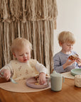 Two light-haired children eat at a wooden table. The younger uses the Mushie Classic Silicone Suction Plate, while the older holds bread. Both have cups nearby, and a textured tapestry is visible in the background.