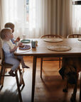 Two young children eat from Mushie Classic Silicone Suction Plates at a dining table, with mugs beside them. A large brown dog stands under the table as sunlight streams through sheer curtains in the background.
