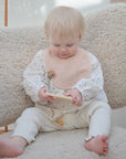 A blond toddler sits on a soft armchair, wearing a pale pink mushie Muslin Bib made from organic cotton and white clothes, looking down while playing with a wooden toy.