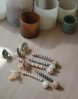 Three mushie Silicone Pacifier Clips | Luna, crafted from food-grade silicone in neutral and pastel colors, are displayed on a wooden surface with candle holders in the background and sunlight casting gentle shadows.