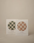 Two boxes of mushie Extra Soft Muslin Changing Pad Covers—one green and white checkered, one pink and white checkered—are shown against a neutral beige background.
