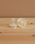 Two cream-colored FRIGG Daisy Silicone Pacifiers with scalloped edges rest on a light wooden surface against a beige background; one stands upright while the other lies flat, showing the back. Comes as a 2-pack from FRIGG and is made from medical grade silicone.