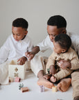 Three young children play on the floor with mushie’s Shape Sorting Box and pastel shape blocks, while two older kids assist a baby in a beige sweater, encouraging hands-on learning and fine motor skills against a neutral background.