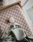 The mushie Extra Soft Muslin Changing Pad Cover in beige and white checks sits by a sunlit window, topped with a green cloth, a small spatula-like tool, and a white bear-shaped object.