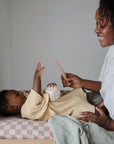 A smiling woman sits beside a baby in yellow, who holds a toy and reaches up while lying on the mushie Extra Soft Muslin Changing Pad Cover, creating a calm, nurturing scene.
