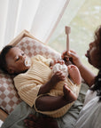 A baby with a pacifier rests on the mushie Extra Soft Muslin Changing Pad Cover atop a checkered pad, while an adult holds a white toy and spatula-like item nearby. Light streams through sheer curtains in the background.