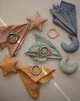 The mushie Star Lovey Blanket is displayed on a light surface with sunlight casting diagonal lines, alongside soft organic cotton cloths in muted colors, star and moon-shaped pillows, and silicone teething rings with cat and bear ear designs.