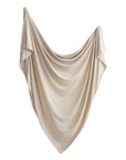 The mushie Stretchy Swaddle, in beige silky fabric, hangs in loose elegant folds with a pointed bottom and higher top corners against a plain white background, evoking the look and feel of a soft baby blanket.