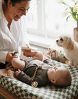 A woman changes her baby’s diaper on a mushie Extra Soft Muslin Changing Pad Cover as a small white dog watches. Natural light fills the room, and fresh flowers decorate the windowsill.