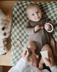 A baby in a brown onesie lies on a green and white checkered changing pad, holding a teething toy. An adult applies lotion with the Mushie Diaper Cream Applicator, while a small white dog sits nearby on the wooden table.