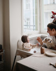 A woman holds a small dog while feeding a baby in a high chair by the window. The baby, wearing a bib and bonnet, enjoys their meal from the Mushie Classic Silicone Suction Plate on the bright, cozy table with dishes and a cup.