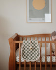 A baby in a knit hat sits in a wooden crib with a polka-dot sheet and mushie Organic Cotton Muslin Burp Cloth 2-Pack nearby. A green and white checkered blanket hangs on the rail, with modern art on the wall behind.