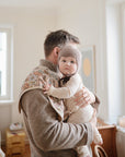 A man in a beige sweater holds a baby in a knit bonnet and cream outfit, with mushie's Organic Cotton Muslin Burp Cloth 2-Pack draped nearby in a cozy, sunlit room with large windows and soft decor.