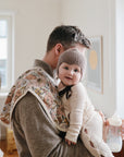 A man holds a smiling baby in a knit bonnet and light outfit, with a mushie Organic Cotton Muslin Burp Cloth over his shoulder and a baby bottle in hand, standing in a bright, cozy room filled with soft natural light.