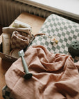 A wooden table holds a mushie Extra Soft Muslin Changing Pad Cover, a peach muslin cloth, a pacifier clip, a green teether, and a small box with baby essentials like lotion, a washcloth, and a pouch.