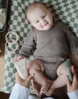 A smiling baby in a brown onesie lies on a green-and-white checkered pad, holding cream while an adult uses the Mushie Diaper Cream Applicator and green spatula. Baby products and toys are on the table nearby.