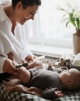 A smiling woman plays with a baby in a grey outfit on a checkered changing pad by the window, surrounded by flowers, baby essentials, and the Mushie Diaper Cream Applicator. The baby happily reaches up toward her.