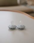 Two FRIGG Daisy Silicone Pacifiers, made from medical grade silicone, with round handles, sit on a white table. This 2-pack by FRIGG offers comfort for babies. The softly blurred background features neutral-toned furniture.