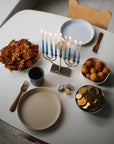 A Hanukkah table with a menorah, mushie Round Dinnerware Plates of latkes, sufganiyot, chocolate gelt, dreidels, a cup, and a bear-shaped child’s chair—all bathed in natural light.