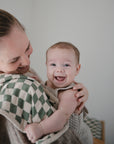 A woman smiles while holding a happy baby indoors, with a light background. She has a mushie Organic Cotton Muslin Burp Cloth from the 2-Pack draped over her shoulder in a green and white checkered pattern.