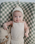 A baby in a cream knit bonnet and sleeveless top smiles while lying on the mushie Extra Soft Muslin Changing Pad Cover, featuring a green and white checkered pattern. A beige cloth and wooden toy rest nearby.
