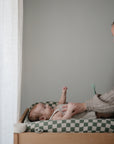A smiling woman leans over a baby lying on a mushie Extra Soft Muslin Changing Pad Cover placed on a checkered pad by a window with sheer curtains, gently holding the baby's hands.