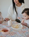 A young child enjoys bananas from the Mushie Classic Silicone Suction Plate with a fork at a table. Another child holds a cup, while an adult sits nearby. A colorful floral tablecloth and a bowl of fruit decorate the table.