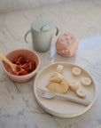 A Mushie Classic Silicone Suction Plate with banana and apple slices, a pink bowl of strawberries, forks and spoons, a green sippy cup, and a pink toy are arranged on a marble surface for a toddler’s snack.