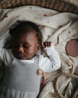 A baby sleeps peacefully in a woven basket lined with the mushie Organic Cotton Muslin Swaddle Blanket, while a round teether toy rests nearby on the cream patterned blanket.