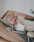 A baby in a white onesie lies on the mushie Extra Soft Muslin Changing Pad Cover, wearing a gray knit hat and raising a fist. A pacifier and jar are nearby as an adult holds a toothbrush with toothpaste.