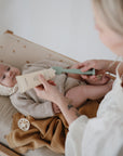 A baby in a knit bonnet and sweater lies on a mushie Extra Soft Muslin Changing Pad Cover as an adult applies lotion to their legs. A small toy and blanket are beside the baby.