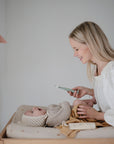 A smiling woman holds a phone while changing her baby—dressed in a knit bonnet and sweater—on the mushie Extra Soft Muslin Changing Pad Cover. Colorful fabric pennants hang in the background as the baby looks up at her.