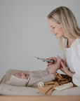 A woman smiles while changing her baby, who’s dressed in a light knit outfit and hat. She gently holds the Mushie Diaper Cream Applicator, ready to use it, as baby care items sit nearby.